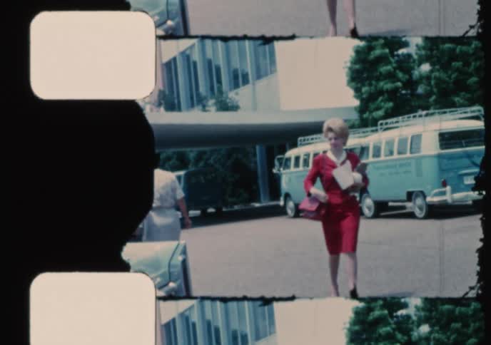 Archival 16mm film shows a stylish woman in a red suit walking past vintage Volkswagen vans in a parking lot during the 1960s