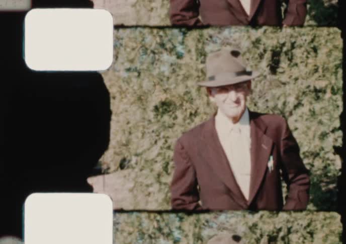 A dapper elderly gentleman in a suit and fedora smiles for the camera in a sunny yard, authentic vintage 8mm home movie footage