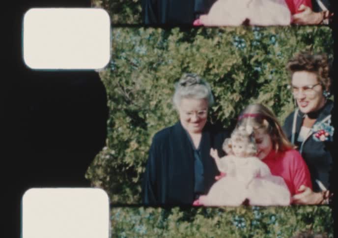 A vintage 16mm film scan shows a happy young girl holding a doll while posing for a family portrait with her mother and grandmother outdoors