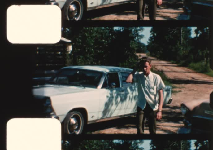 Archival 16mm film scan of a young man posing with his vintage car on a rural road in the 1960s