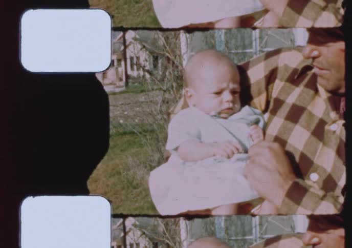 A father or grandfather in a plaid shirt and hat gently holds a small baby outdoors in a yard on a sunny day