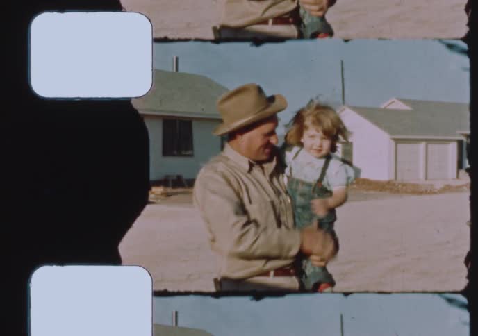 A proud father in a hat smiles and picks up his young child in front of their house in this vintage home movie