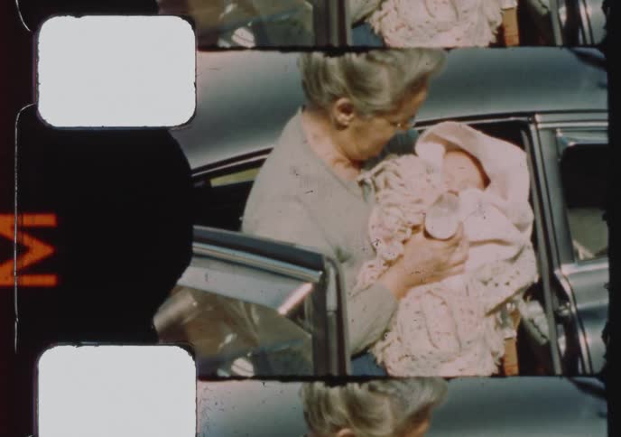 A happy grandmother lovingly holds her newborn grandchild next to a vintage car in this archival 8mm film