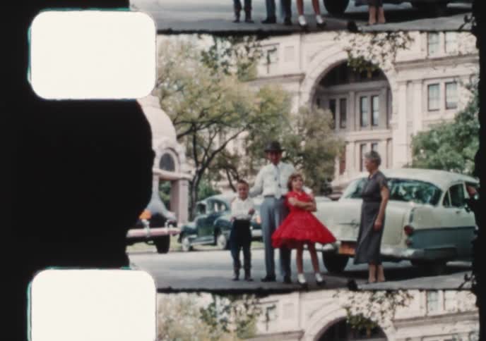 A multi-generational family in 1950s attire poses for a photo in a town square surrounded by vintage cars