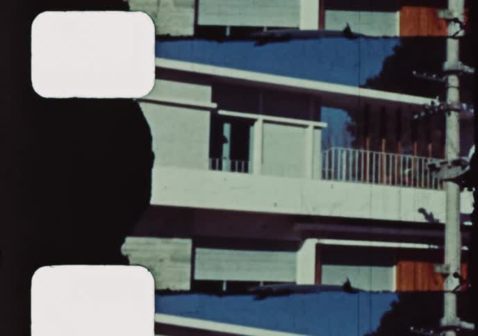 Vintage 8mm footage pans from a classic car on the street up to a woman waving from a modern apartment balcony