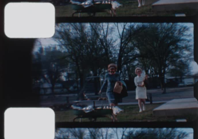 Two stylish women from the 1960s walk across a suburban front yard carrying bags on a cloudy day