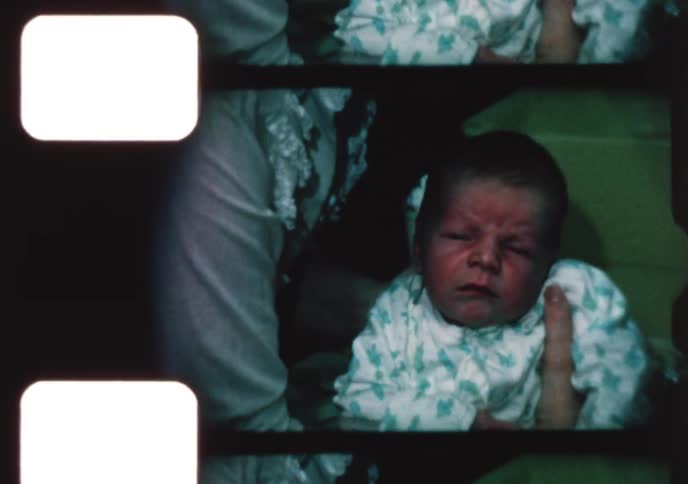 A vintage home movie captures an intimate close-up of a newborn baby with a serious expression being held and comforted by their mother
