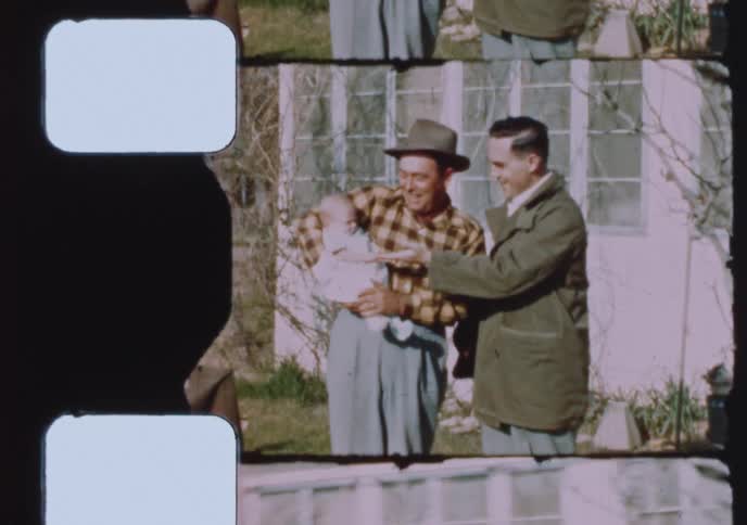 Vintage 8mm film scan shows two smiling men admiring a newborn baby outside a house