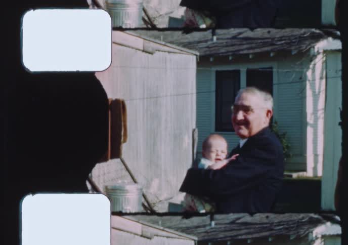 A vintage 8mm film scan captures a tender moment as a proud grandfather holds his newborn grandchild in the backyard of a home