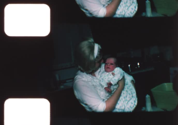 A new mother tenderly holds her crying newborn baby after giving it a bath with help from another woman in a vintage 1960s home movie