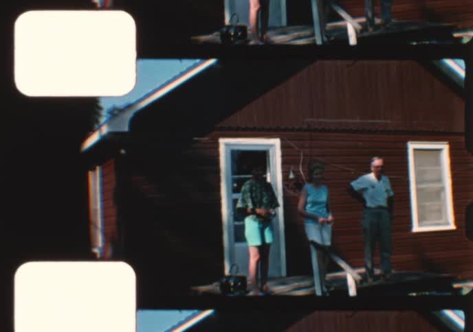 A mother helps her toddler learn to walk in the sunny yard of a rustic cabin during a family gathering in this authentic vintage home movie