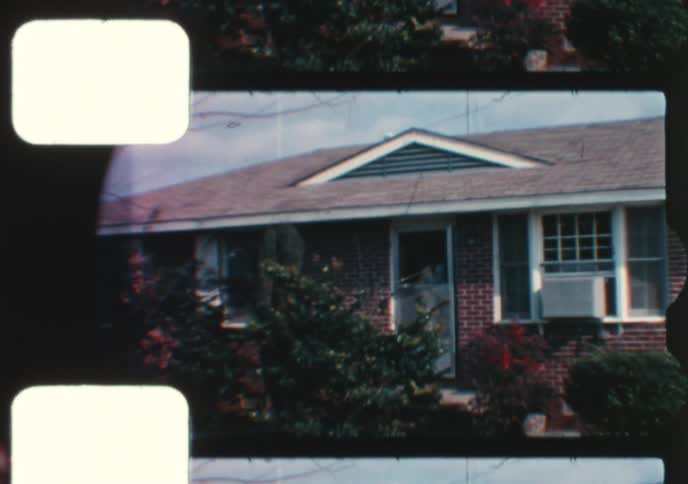 A vintage 8mm home movie captures a mother holding her baby outside their brick suburban home
