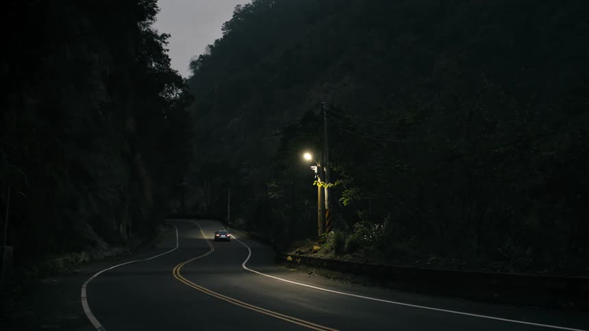Two cars drive down a lonely winding mountain road illuminated by a single street light at night