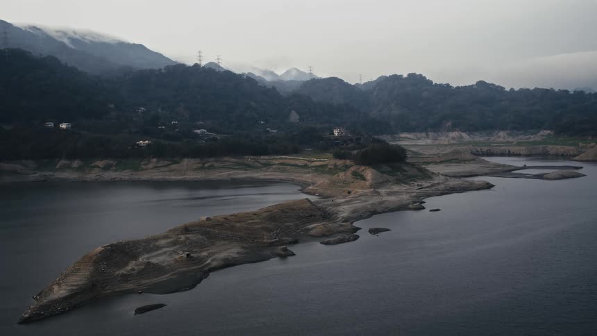 A desolate, wide view shows a drought-stricken reservoir with critically low water levels surrounded by misty mountains