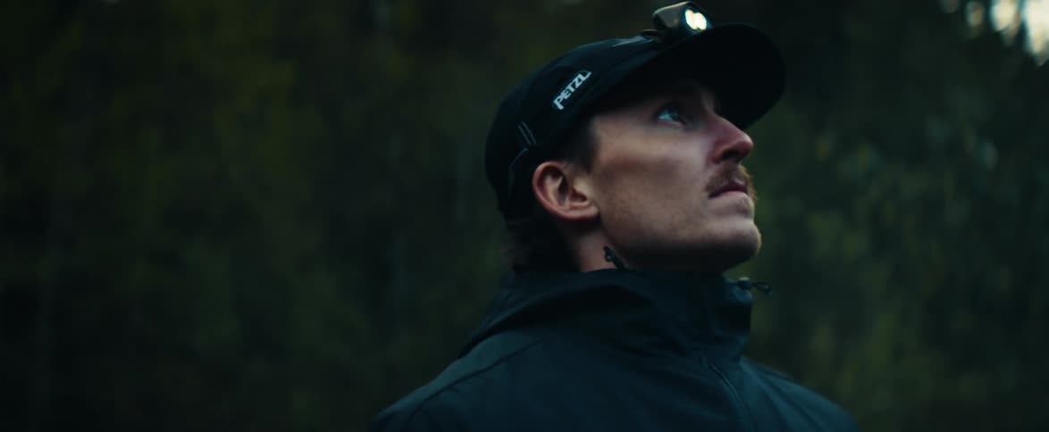 A hiker wearing a headlamp looks up into the dark forest canopy, observing his surroundings before moving on