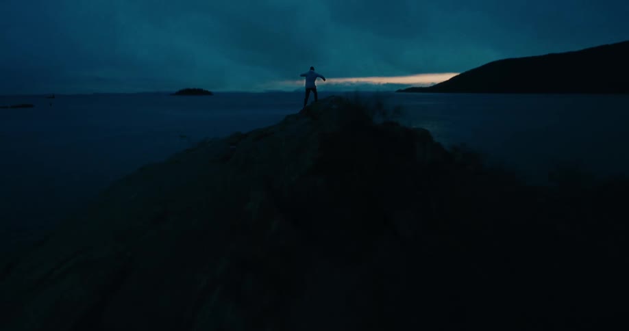 An adventurous person runs to the summit of a rocky island and raises their arms in triumph as a powerful lightning storm illuminates the dark night sky