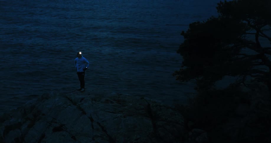 A solitary trail runner with a headlamp pauses on a rocky cliff to look out at the dark ocean waves at night