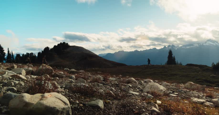 A silhouetted hiker traverses a rocky alpine landscape with a vast mountain range in the background as the sun shines through the clouds