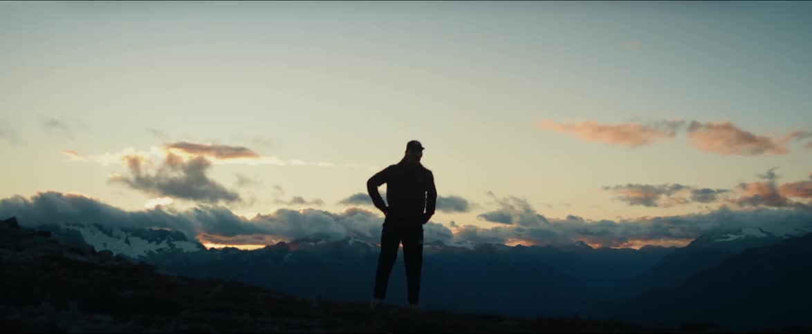 A lone trail runner's silhouette is captured in slow motion as he conquers a mountain ridge against a dramatic sunset and snowy peaks