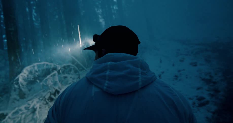 Over-the-shoulder shot of a person running through a dark and snowy forest at night, their headlamp cutting through the gloom as snow falls