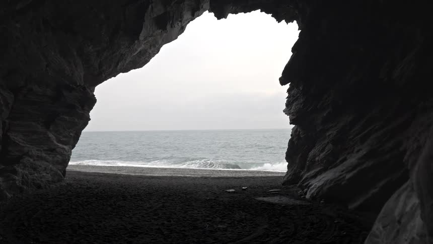 A dramatic view from inside a dark rock cave looking out at the ocean waves rolling onto a black sand beach on an overcast day