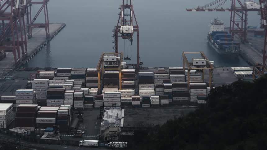 A large crane moves shipping containers around a busy commercial port on a gray, overcast day