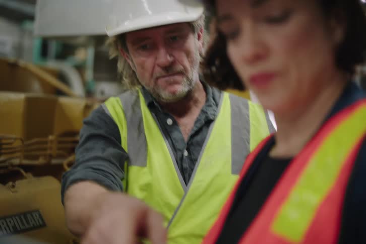 A male mechanic instructs a female colleague on heavy machinery while she uses a laptop in a workshop