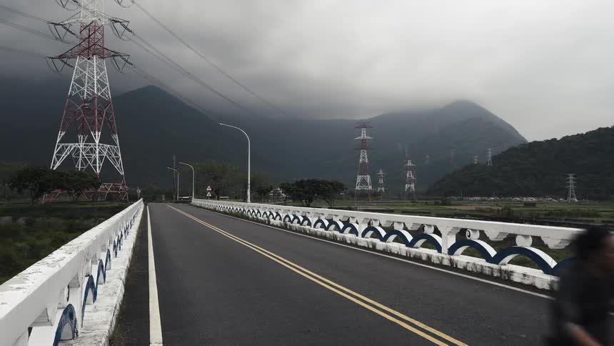 A man on a touring bicycle rides towards the camera across a bridge in a misty mountain valley