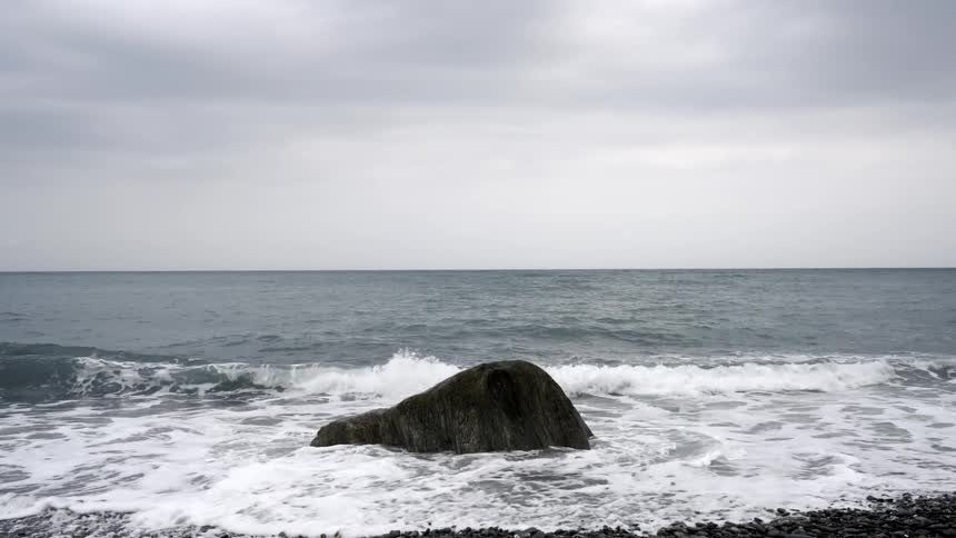 Slow motion shot of ocean waves washing over a large rock on a pebbled beach on an overcast day