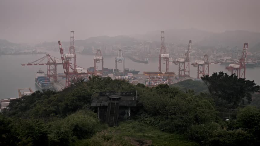 An old, derelict bunker on a lush hillside overlooks a large, industrial shipping port on a foggy day