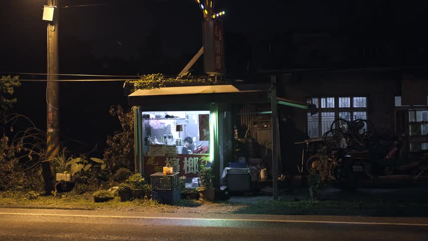 A lone food vendor works inside a small, illuminated roadside kiosk on a dark and quiet night in rural Asia