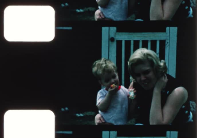 Vintage home movie from the 1970s shows a young mother and her toddler son enjoying an Easter egg hunt in their sunny backyard