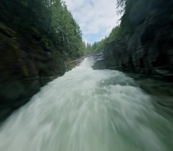 An exhilarating FPV drone flight speeds up a rushing river through a narrow rocky canyon towards a cascading waterfall