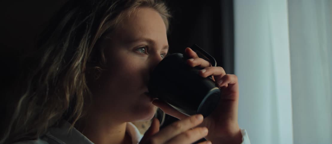A young blonde woman in a white shirt thoughtfully sips coffee from a black mug while looking out a window in slow motion