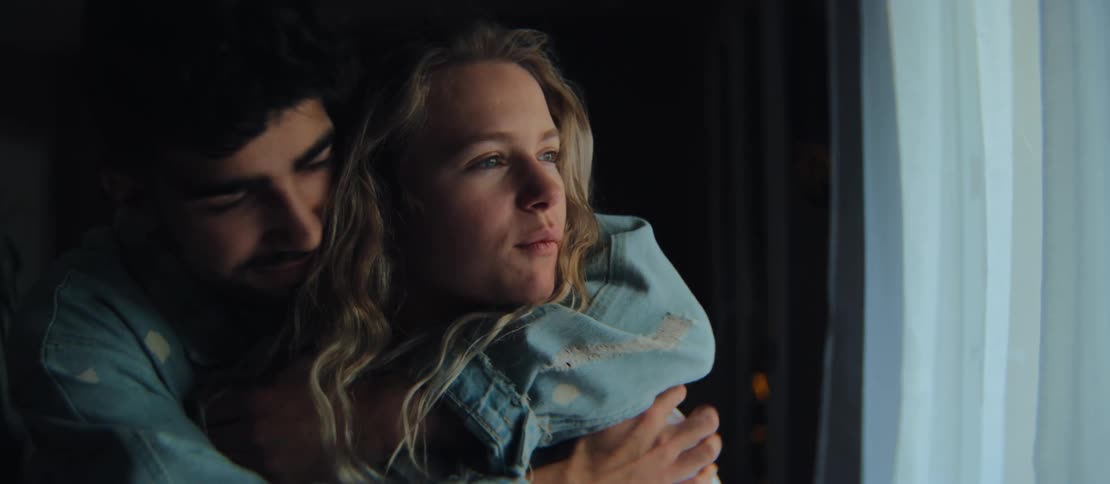 A young, romantic couple shares a tender embrace and kiss by a window at night, enjoying a quiet, intimate moment together at home