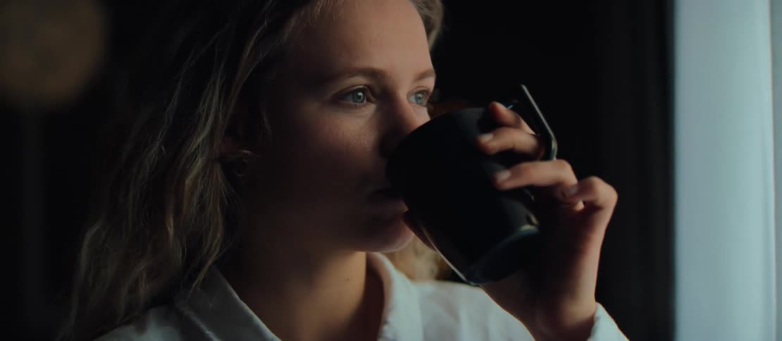A contemplative young woman with blonde hair slowly sips from a black mug in a dimly lit room, gazing into the distance