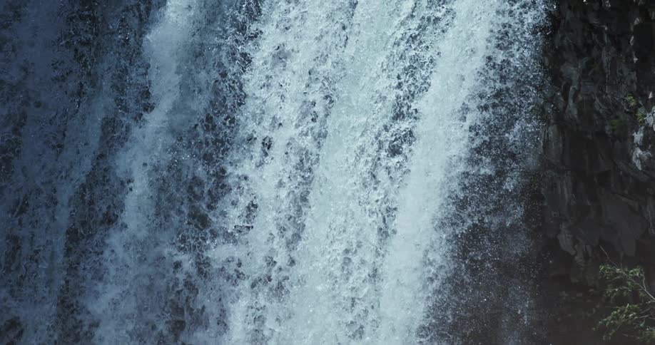 A dramatic slow-motion shot tilts up a powerful waterfall, revealing the river cascading over a rocky cliff