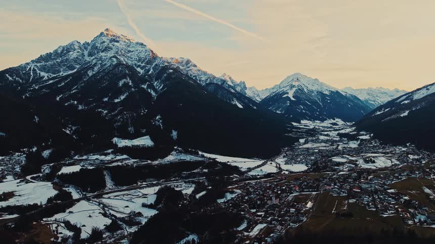A serene aerial view shows a charming mountain town nestled in a snow-covered valley as the setting sun illuminates the majestic peaks
