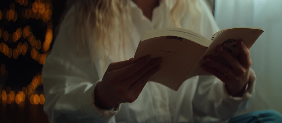 A woman with blonde hair reads a book in a dimly lit, cozy room with soft lights in the background