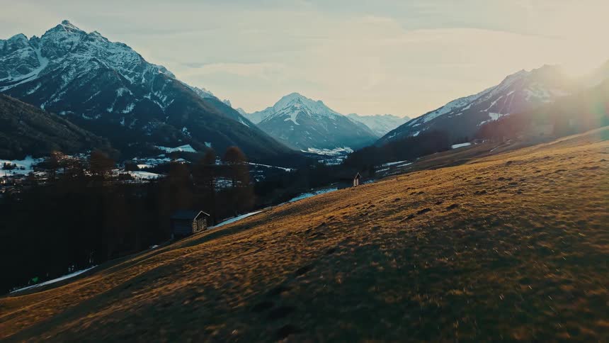 An aerial view flies past a rustic wooden cabin on a golden hillside at sunset, revealing a stunning snow-capped mountain valley