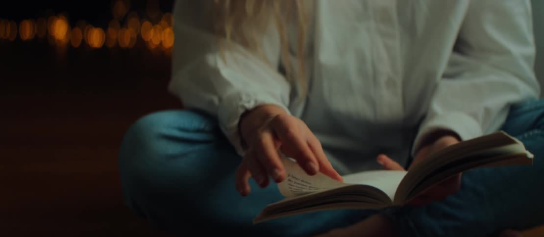 A woman's hands gently turn the pages of a book as she reads, her fingers tracing the words in a cozy, dimly lit room