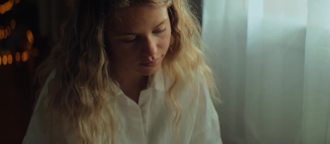 A cinematic slow-motion shot of a thoughtful young woman with curly blonde hair engrossed in reading a book indoors