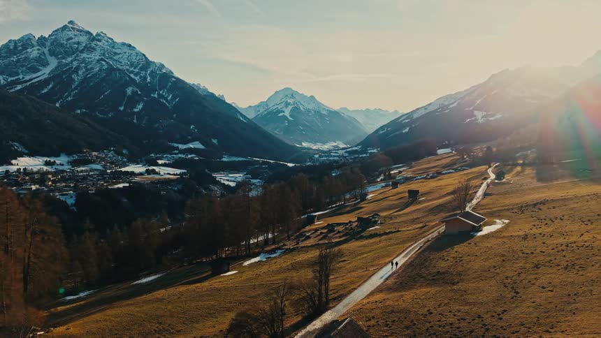 An epic aerial view shows two people walking along a scenic path through a golden valley surrounded by snow-capped mountains at sunset
