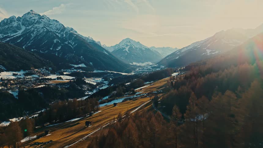 A majestic aerial flight over a forested hillside reveals a vast, sunlit valley surrounded by snow-capped mountains