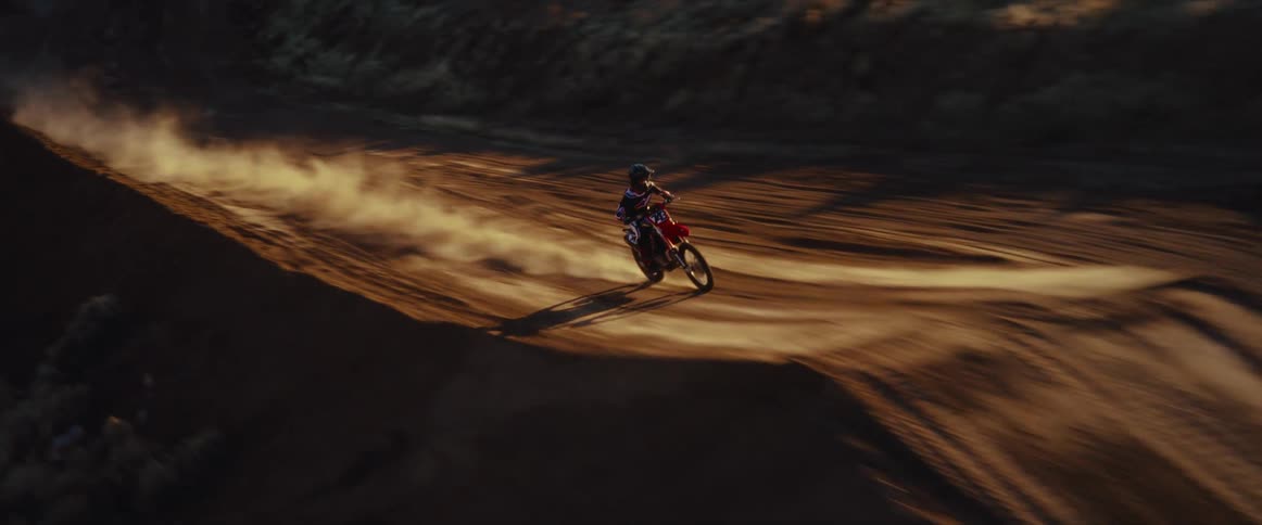 A motocross rider races around a dirt track and launches off a jump in dramatic slow motion, captured by a drone at sunset