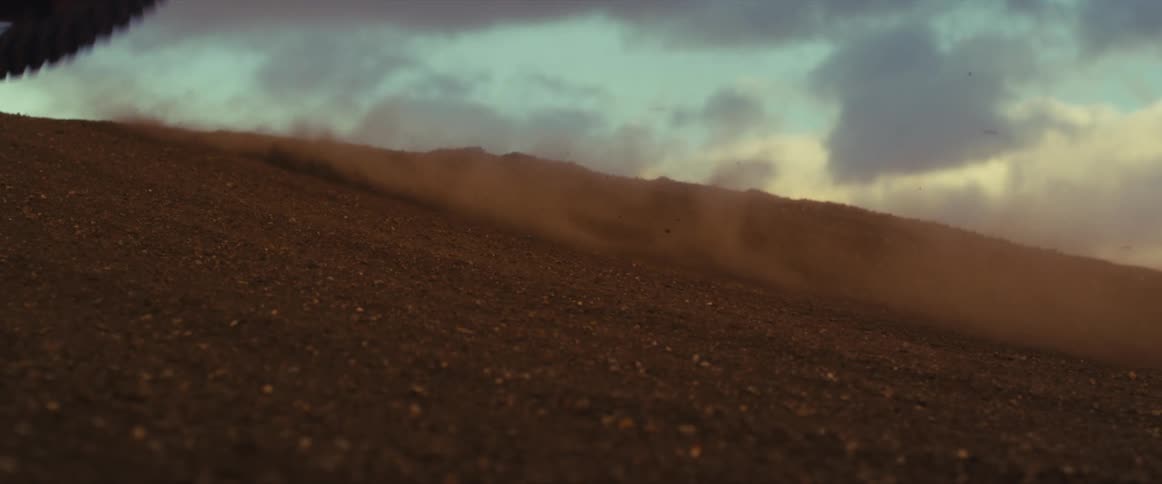 A motocross bike tire spins in slow motion as it climbs a dirt hill, kicking up a cloud of dust at sunset