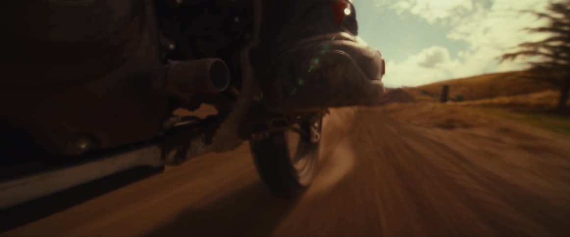 A slow-motion close-up shows a motocross bike jumping over a dirt mound, with the sun flaring behind the rider's boot