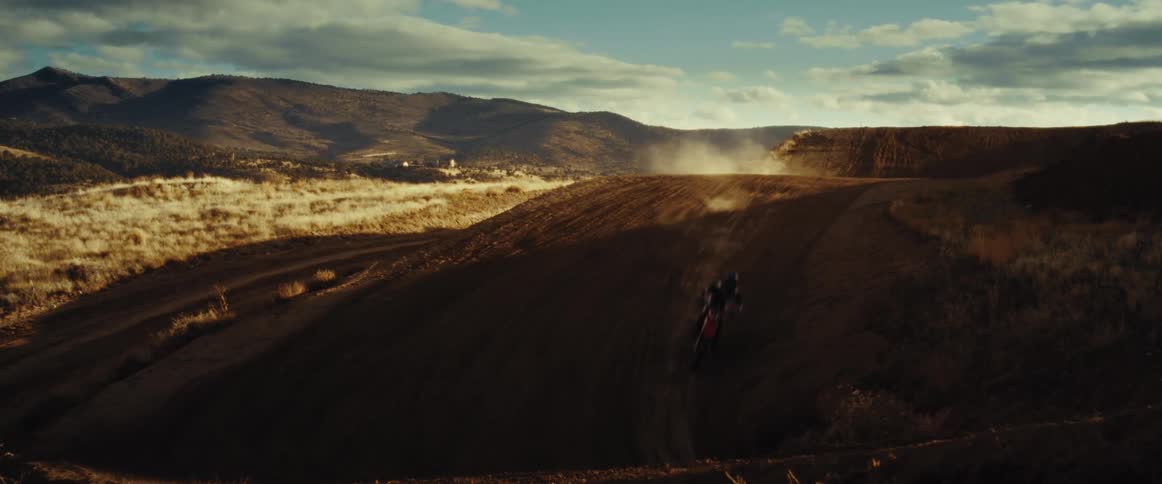 A motocross rider lands a big jump and speeds down a dusty dirt track in a rugged landscape during golden hour