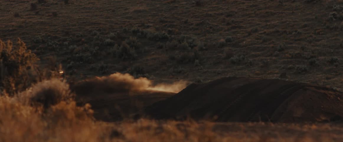 Motocross rider speeds over a dirt jump in slow motion, kicking up a large cloud of dust during golden hour