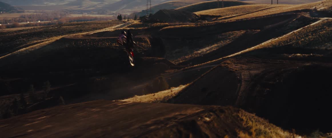 Slow-motion aerial shot of a motocross rider performing a big air jump over a winding dirt track in the golden hills